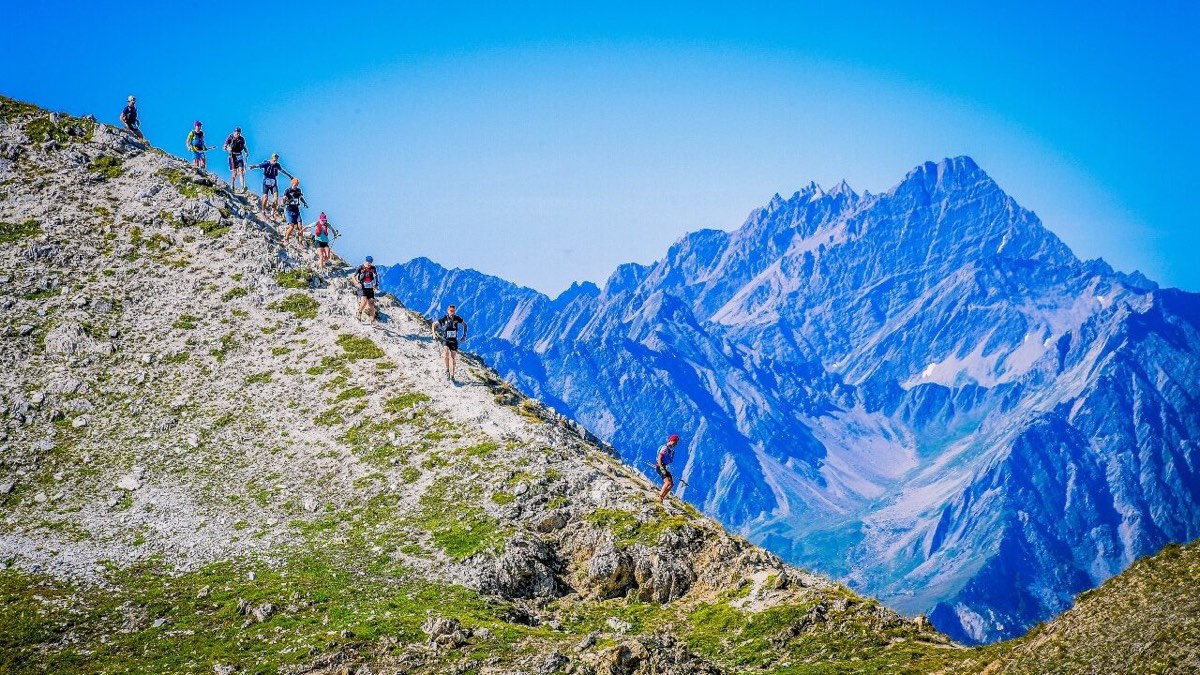 Trail runners descending a rocky alpine ridge with jagged Alps peaks behind