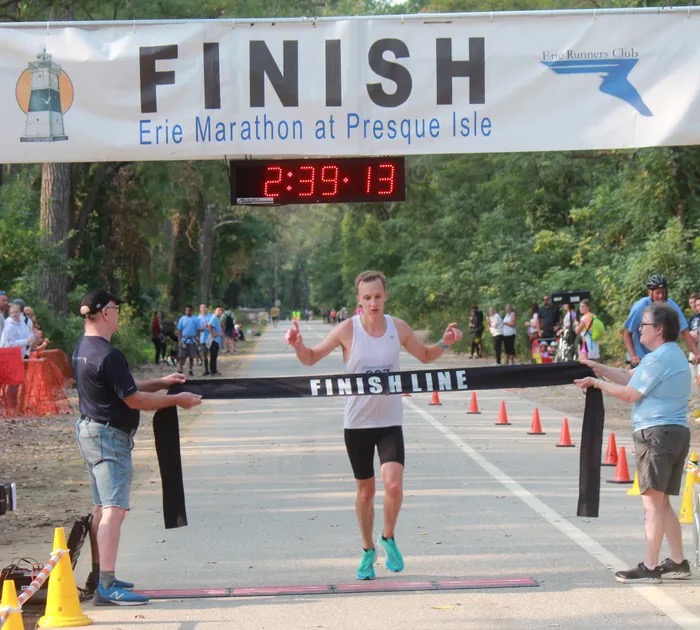 A runner breaking the finish tape under the Erie Marathon at Presque Isle finish banner