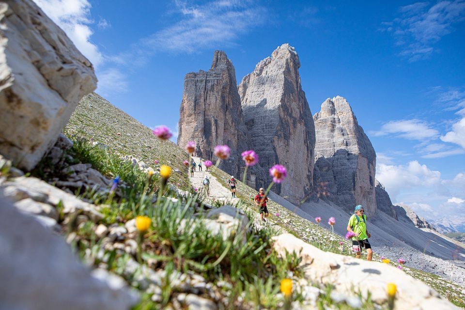 Lavaredo Ultra Trail runners on a wildflower-lined trail beneath the Tre Cime di Lavaredo in the Dolomites