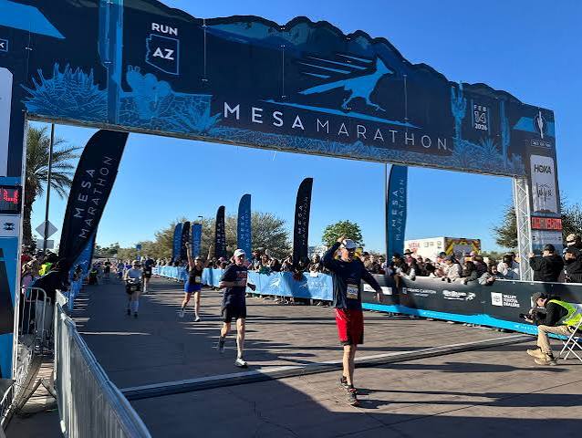 Runners crossing the Mesa Marathon finish line under a blue arch on a sunny day